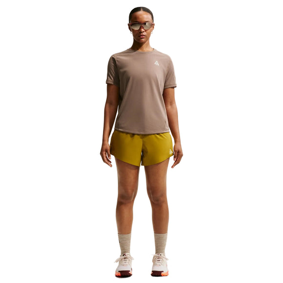 En person ser fremad, iført solbriller, Nike ACG Short-Sleeve Running Top til kvinder i Mink Brown/Summit White, gule shorts, grå sokker og hvide sportssko, med armene afslappet langs siden mod en almindelig hvid baggrund.