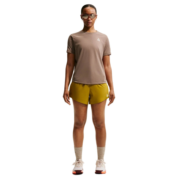 En person ser fremad, iført solbriller, Nike ACG Short-Sleeve Running Top til kvinder i Mink Brown/Summit White, gule shorts, grå sokker og hvide sportssko, med armene afslappet langs siden mod en almindelig hvid baggrund.