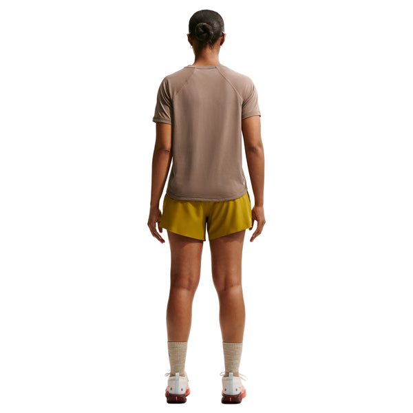 En person kigger væk fra kameraet i en Nike ACG kortærmet løbetop til kvinder, Mink Brown/Summit White, sammen med gule shorts, lyse strømper og hvide sko - ideelt til trailløb. Baggrunden er hvid.