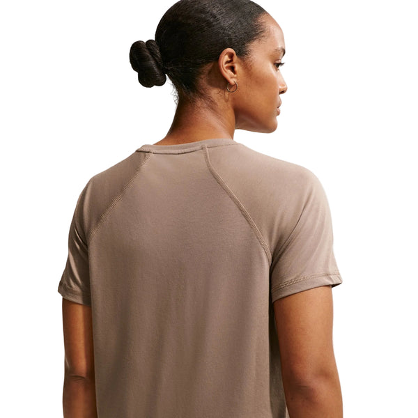 En person med mørkt hår i en lav knold ses bagfra iført Nike ACG Short-Sleeve Running Top til kvinder i Mink Brown/Summit White. Baggrunden er hvid.