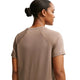 En person med mørkt hår i en lav knold ses bagfra iført Nike ACG Short-Sleeve Running Top til kvinder i Mink Brown/Summit White. Baggrunden er hvid.
