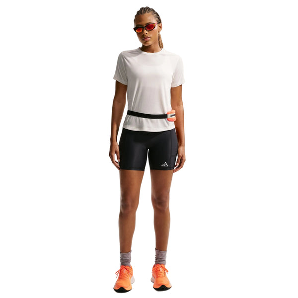 En person står med ansigtet fremad i en Nike ACG kortærmet løbetop til kvinder (Phantom/Summit White), sorte tætsiddende shorts, grå sokker, orange løbesko, røde solbriller og en lille taljetaske - klar til trailløb på en hvid baggrund.