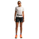 En person står med ansigtet fremad i en Nike ACG kortærmet løbetop til kvinder (Phantom/Summit White), sorte tætsiddende shorts, grå sokker, orange løbesko, røde solbriller og en lille taljetaske - klar til trailløb på en hvid baggrund.