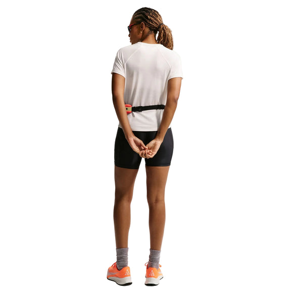 Set bagfra står en person med foldede hænder iført Nikes ACG Short-Sleeve Running Top til kvinder i Phantom/Summit White - sammen med sorte shorts, grå sokker, orange sko, solbriller og et bælte - klar til trailløb.