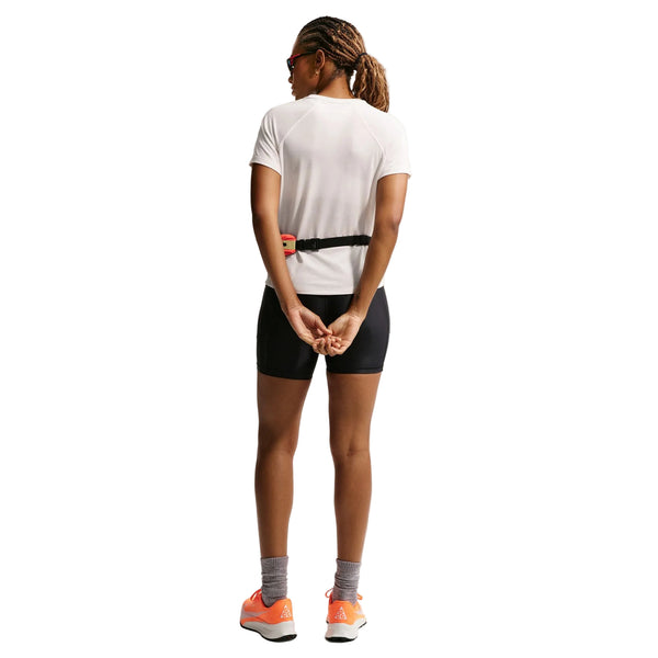 Set bagfra står en person med foldede hænder iført Nikes ACG Short-Sleeve Running Top til kvinder i Phantom/Summit White - sammen med sorte shorts, grå sokker, orange sko, solbriller og et bælte - klar til trailløb.