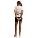 Set bagfra står en person med foldede hænder iført Nikes ACG Short-Sleeve Running Top til kvinder i Phantom/Summit White - sammen med sorte shorts, grå sokker, orange sko, solbriller og et bælte - klar til trailløb.