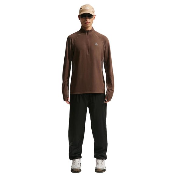 En person står med front mod en almindelig hvid baggrund iført en beige kasket, en brun langærmet skjorte med lynlås, Nike ACG Trailwind Waterproof Pants til mænd i Black/Summit White og hvide sneakers.