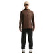 En person står med front mod en almindelig hvid baggrund iført en beige kasket, en brun langærmet skjorte med lynlås, Nike ACG Trailwind Waterproof Pants til mænd i Black/Summit White og hvide sneakers.
