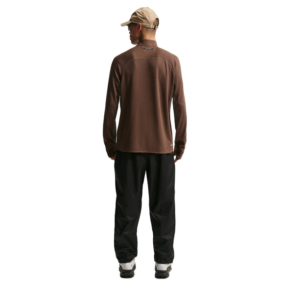 En person iført beige kasket, brun langærmet skjorte og Nike ACG Trailwind Waterproof Pants til mænd i Black/Summit White står med ansigtet væk fra kameraet mod en almindelig hvid baggrund.