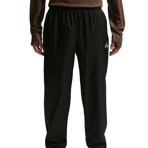 En person iført løstsiddende Nike ACG Trailwind Waterproof Pants til mænd i Black/Summit White, med elastisk linning og et lille hvidt logo på venstre lår, sammen med en langærmet brun top. Billedet er beskåret ved skuldre og ankler.