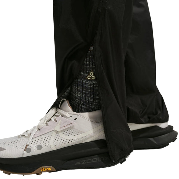 Nærbillede af en person iført hvide Nike-sneakers med sorte såler, strømper med sort og hvidt ACG-logo og Nike ACG Trailwind Waterproof Pants til mænd i Black/Summit White.