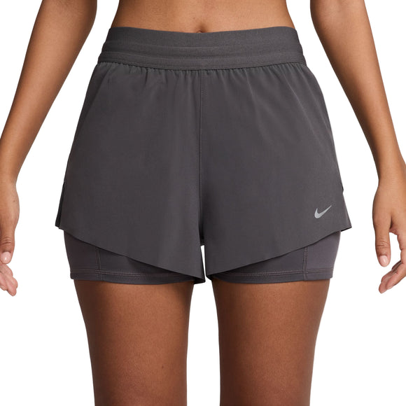 En person står med afslappede arme iført Nike Dri-FIT Mid-Rise 2-i-1 løbeshorts til kvinder i Thunder Grey/Reflective Silver. Nike-logoet ses nederst i venstre side af shortsens lagdelte design.