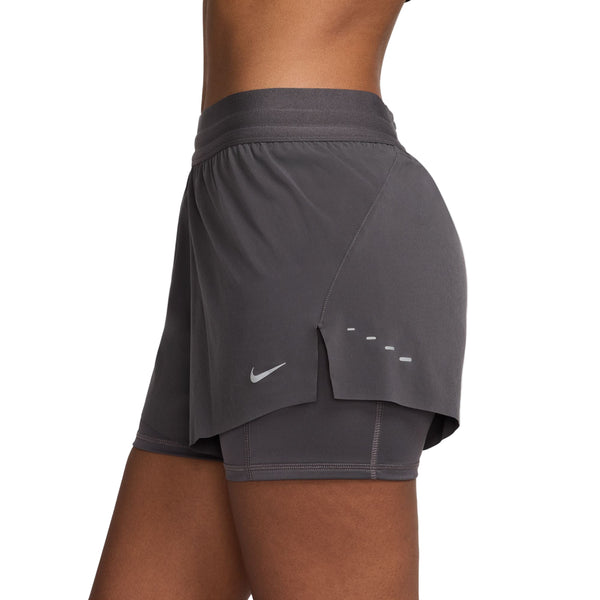 En person står med siden til kameraet iført Nike Dri-FIT Mid-Rise 2-i-1 løbeshorts til kvinder i Thunder Grey/Reflective Silver på en almindelig hvid baggrund.