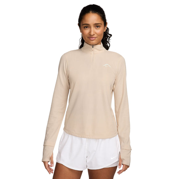 En kvinde med mørkt bølget hår, der er trukket tilbage, bærer Nike Dri-FIT Trail 1/2-Zip Mid Layer til kvinder i Pearl White/Summit White og hvide sportsshorts, mens hun kigger fremad med et neutralt udtryk mod en almindelig hvid baggrund.