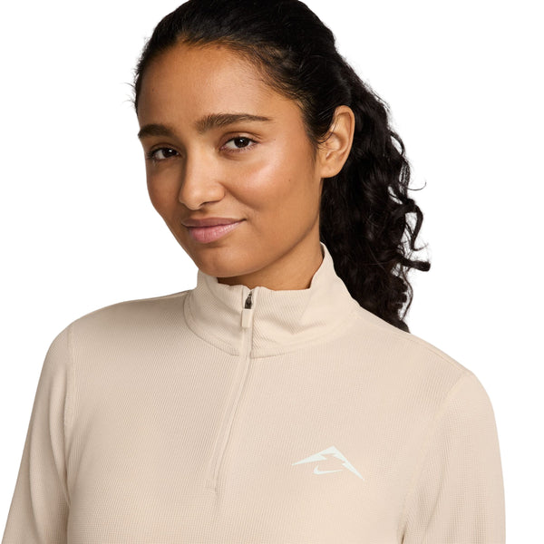 En person med langt, krøllet mørkt hår er iført en lys beige Nike Dri-FIT Trail 1/2-Zip Mid Layer til kvinder i Pearl White/Summit White med et lille hvidt logo på brystet, der står på en almindelig hvid baggrund.