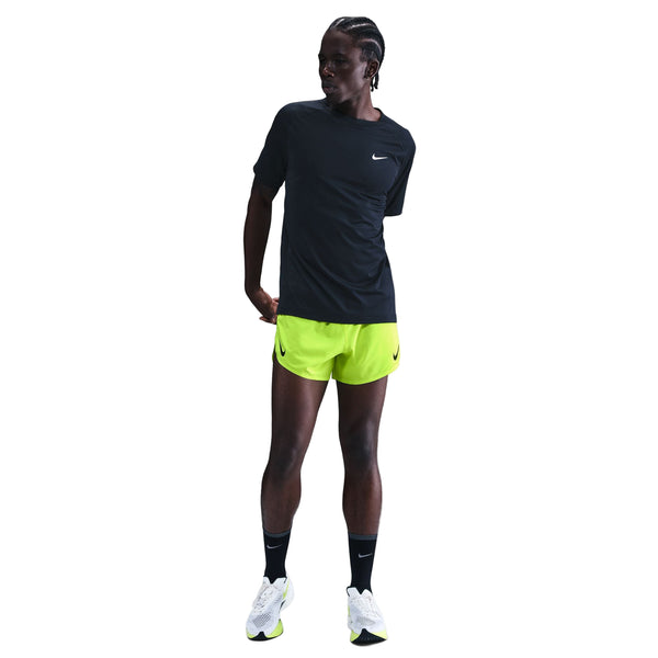 En person i sort Nike t-shirt, sorte knæstrømper, hvide sportssko og Nike Dri-FIT ADV 4" Brief-Lined Running Shorts for men (Volt/Black) står på en hvid baggrund og kigger til siden med den ene arm bag ryggen.