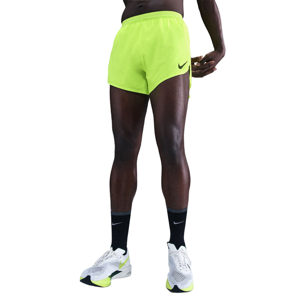En mand i Nike Dri-FIT ADV 4" Brief-Lined Running Shorts (Herre, Volt/Black) står på en hvid baggrund sammen med hvide og neongrønne Nike-løbesko og sorte Nike-sokker.
