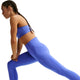 En kvinde i Nike Nk Df Swift Hr 7/8 Tight, Dame, Sapphire/Reflective Silv og en matchende sports-bh laver udfald med hænderne på sit bøjede knæ. Disse åndbare leggings har Nike Dri-FIT-teknologi for maksimal komfort under træning, isoleret på hvid baggrund.
