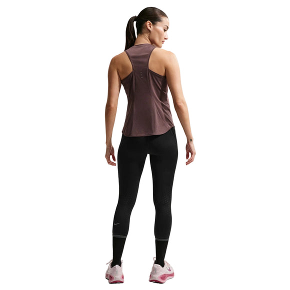 En kvinde med hestehale står med ryggen til og er iført en Nike Nk Swift Df Tank Top til kvinder i Tattoo/Reflective Silv, sammen med sorte leggings og hvide sko med pink såler; hendes arme er afslappede i siden mod en hvid baggrund.