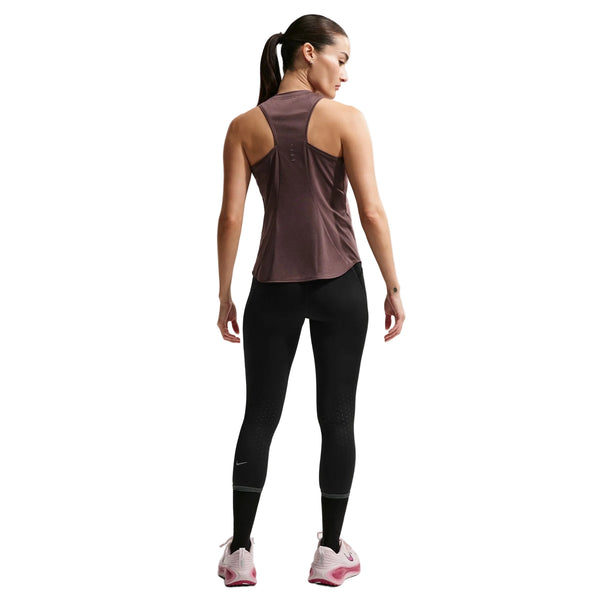 En kvinde med hestehale står med ryggen til og er iført en Nike Nk Swift Df Tank Top til kvinder i Tattoo/Reflective Silv, sammen med sorte leggings og hvide sko med pink såler; hendes arme er afslappede i siden mod en hvid baggrund.