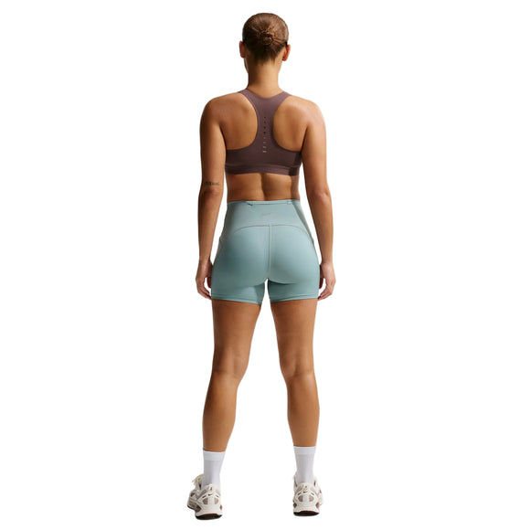 En person står med ryggen til kameraet og har en Nike Nk Swift Hs Bra til kvinder i farven Tattoo/Silver på, lyseblå shorts med høj talje, hvide strømper og hvide sportssko på, foran en ensfarvet hvid baggrund.