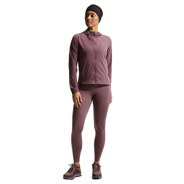 En kvinde bærer en Nike Nk Swift Rpl Pkbl Jkt Dame i Tattoo/Reflective Silv med leggings, sneakers og et sort pandebånd. Jakken med lynlås har åndbarhed og reflekterende detaljer. Hun står på en almindelig hvid baggrund med ansigtet lidt til siden.