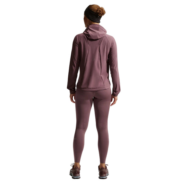 En kvinde kigger væk fra kameraet iført Nike Nk Swift Rpl Pkbl Jkt (Tattoo/Reflective Silv) med matchende leggings, løbesko og et sort pandebånd. Den almindelige hvide baggrund fremhæver hendes sporty look.