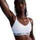 En kvinde med flettet hår bærer en Nike Padded Adjustable Sports Bra til kvinder i hvid/sort med Dri-FIT-materiale og et sort logobånd og løfter den ene arm mod en almindelig hvid baggrund.