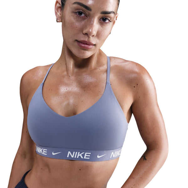 En kvinde ser frem for sig i en Nike Padded Adjustable Sports Bra, Dame, World Indigo, med sorte underbukser. Hendes våde hår er trukket tilbage, og hun er synligt svedig, hvilket tyder på en træning. Baggrunden er helt hvid.