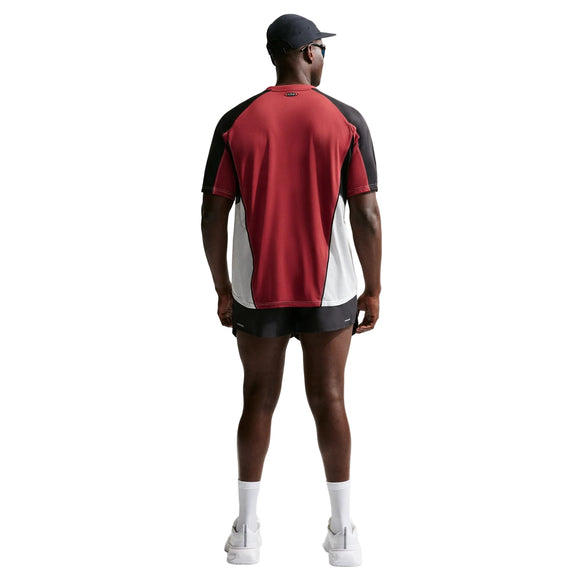 En mand står med ryggen til kameraet iført en Nike Retro SS-løbetrøje i farverne Team Crimson/Black/Reflective Silv, sorte shorts, hvide sokker og sneakers samt en sort kasket foran en ensfarvet hvid baggrund.