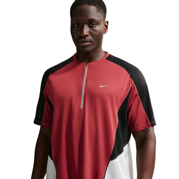 En mand står foran en hvid baggrund iført en Nike Retro SS-løbetrøje til mænd i farvekombinationen Team Crimson/sort/reflekterende sølv.