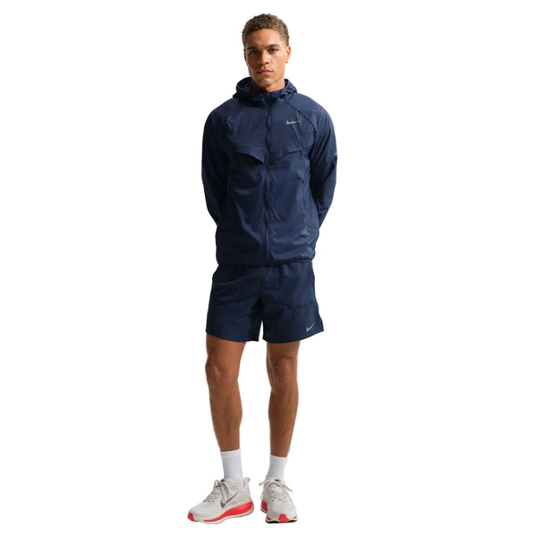 En mand står og kigger fremad i en Nike Stride Jacket, Herre, Midnight Navy/Reflective Silv med matchende shorts, hvide strømper og hvide sneakers med røde detaljer på en almindelig hvid baggrund. Ideel til sporty stil.