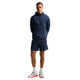 En mand står og kigger fremad i en Nike Stride Jacket, Herre, Midnight Navy/Reflective Silv med matchende shorts, hvide strømper og hvide sneakers med røde detaljer på en almindelig hvid baggrund. Ideel til sporty stil.