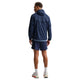 En mand kigger væk fra kameraet iført en Nike Stride Jacket, Herre, Midnight Navy/Reflective Silv. Den herre løbejakke skiller sig ud fra den almindelige hvide baggrund, sammen med marineblå shorts, hvide strømper og hvide sko med røde detaljer.