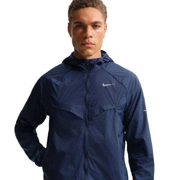 En person iført Nike Stride Jacket, Herre, Midnight Navy/Reflective Silv med lynlås foran står på en almindelig hvid baggrund og ser lige frem.