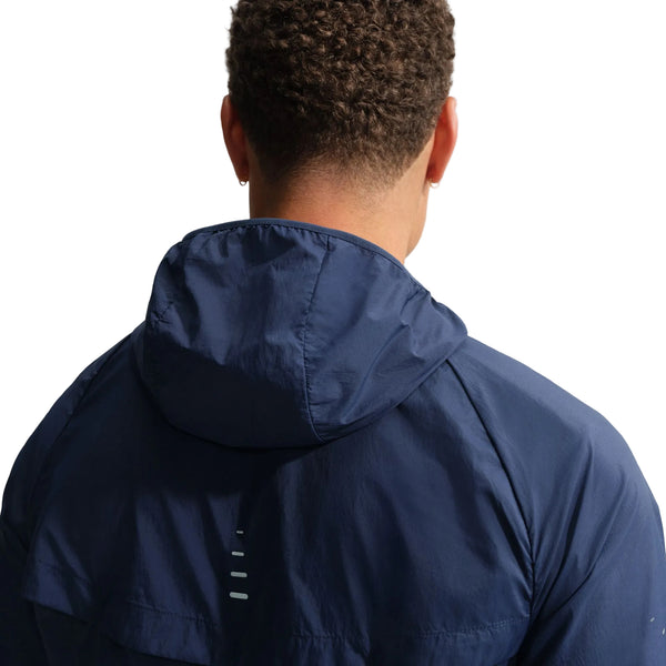 En person med kort, krøllet hår og små øreringe ses bagfra iført Nike Stride Jacket, Herre, Midnight Navy/Reflective Silv. Jakken har reflekterende striber på ryggen. Baggrunden er hvid.