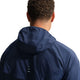 En person med kort, krøllet hår og små øreringe ses bagfra iført Nike Stride Jacket, Herre, Midnight Navy/Reflective Silv. Jakken har reflekterende striber på ryggen. Baggrunden er hvid.