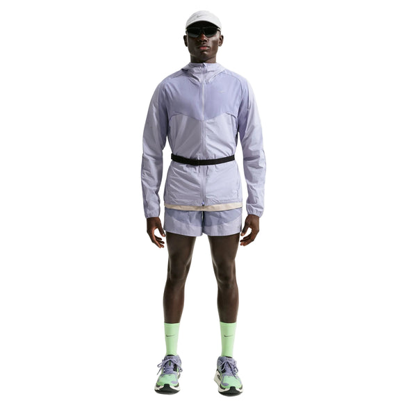 En person står med front mod en hvid baggrund iført en Nike Stride Plus Reple løbejakke til mænd i Iron Purple/Indigo Haze/Reflective Silver, matchende shorts, neongrønne sokker, flerfarvede sneakers, en hvid kasket og solbriller.