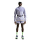 En person står med front mod en hvid baggrund iført en Nike Stride Plus Reple løbejakke til mænd i Iron Purple/Indigo Haze/Reflective Silver, matchende shorts, neongrønne sokker, flerfarvede sneakers, en hvid kasket og solbriller.