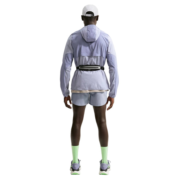 En person kigger væk og er iført en Nike Stride Plus Reple Running Jacket til mænd i Iron Purple/Indigo Haze/Reflective Silver med matchende shorts, neongrønne sokker, hvide løbesko, en hvid kasket og en sort taljetaske på en hvid baggrund.