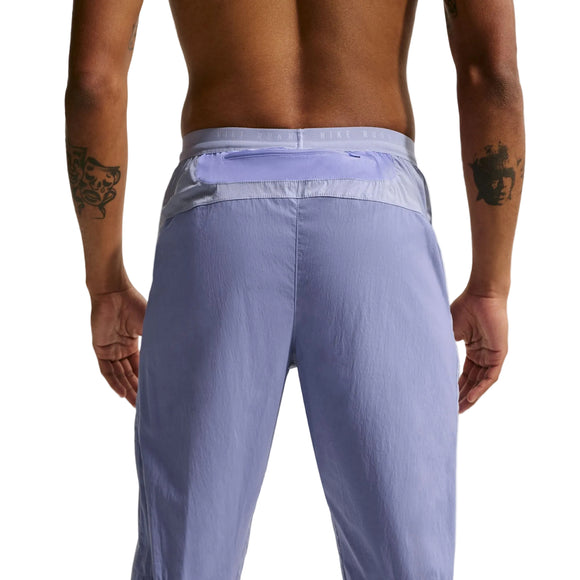 En skjorteløs person med tatoveringer står med ryggen til kameraet iført Nike Stride Repel Running Pants til herrer i Iron Purple/Indigo Haze/Reflective, med lynlåslomme i linningen og atletisk stil.