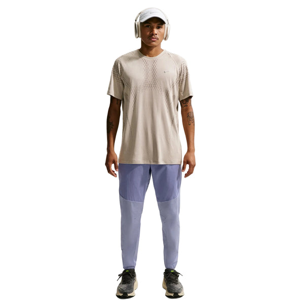 En person iført en beige t-shirt, Nike Stride Repel løbebukser til mænd i Iron Purple/Indigo Haze/Reflective, sorte sneakers, en hvid kasket og hvide hovedtelefoner står med front mod en hvid baggrund.