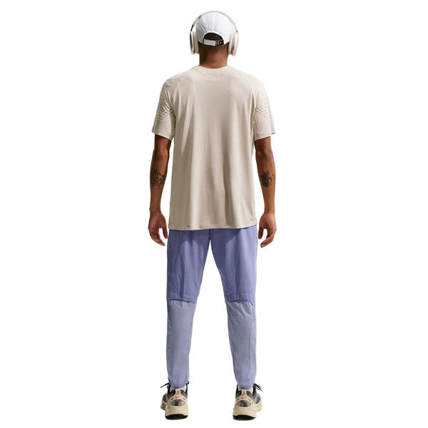 En person står med front mod kameraet iført en beige t-shirt, Nike Stride Repel løbebukser til mænd i Iron Purple/Indigo Haze/Reflective, hvide sneakers, hovedtelefoner og en beige kasket. Der er synlige tatoveringer på begge underarme.