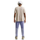 En person står med front mod kameraet iført en beige t-shirt, Nike Stride Repel løbebukser til mænd i Iron Purple/Indigo Haze/Reflective, hvide sneakers, hovedtelefoner og en beige kasket. Der er synlige tatoveringer på begge underarme.