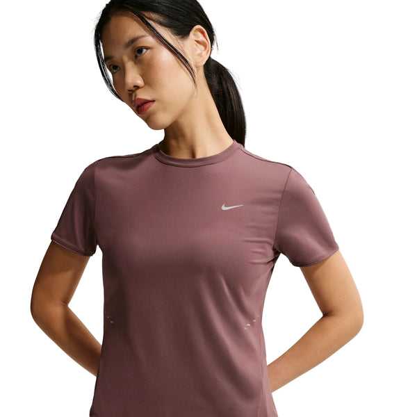 En kvinde med glat sort hår i en lav hestehale står med hænderne på ryggen iført en Nike Swift SS løbetop til kvinder i Tattoo/Reflective Silver med et lille hvidt Nike-logo på venstre bryst.