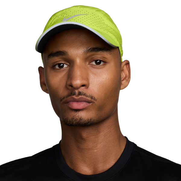 En person i sort skjorte og en lys Cyber/Anthracite/Reflective Silv Nike Unstructured Reflective Cap (unisex) kigger ind i kameraet mod en almindelig hvid baggrund, der fremhæver åndbar kasket-designet.