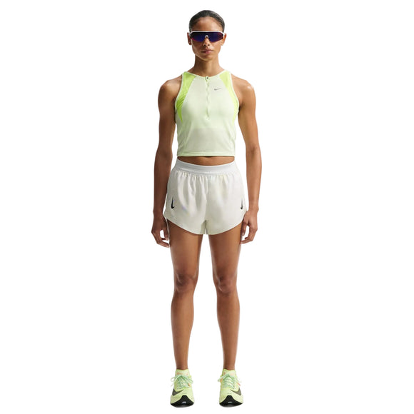 En kvinde kigger fremad iført en lysegrøn ærmeløs top, Nike AeroSwift 3In løbeshorts til kvinder i off white/hvid/sort, grønne løbesko og store reflekterende solbriller mod en hvid baggrund.