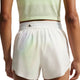 En person ses bagfra iført en lysegrøn ærmeløs top og højtaljede Nike AeroSwift 3In løbeshorts til kvinder i Off White/White/Black, fremstillet i åndbart materiale og med en lille lynlåslomme i linningen.