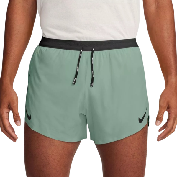 En person iført hvid skjorte og Nike AeroSwift 4" løbeshorts, Herre i Steam/Black, med sort linning, snore og sorte logoer. De svedtransporterende shorts giver komfort, når hænderne hviler i siden.