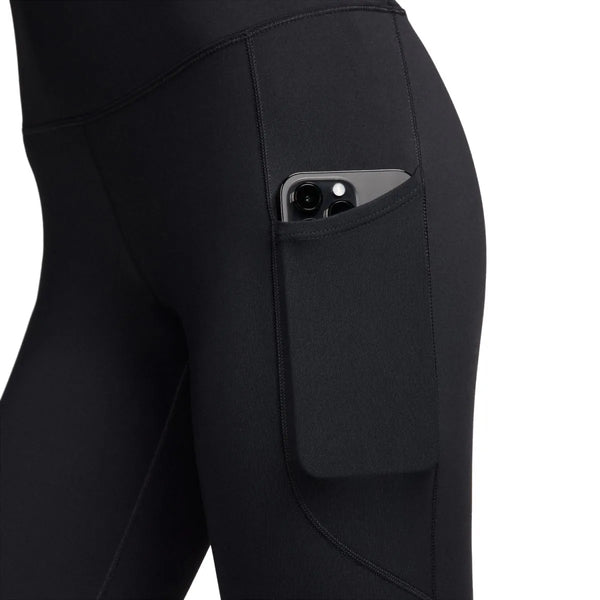Et nærbillede af en person, der har Nike One High-Waisted 7/8, Dame, Black/Black-træningstights på, med en smartphone i sidelommen. Nike-træningstightsene er udstyret med Dri-FIT-teknologi og er fotograferet foran en ensfarvet hvid baggrund.